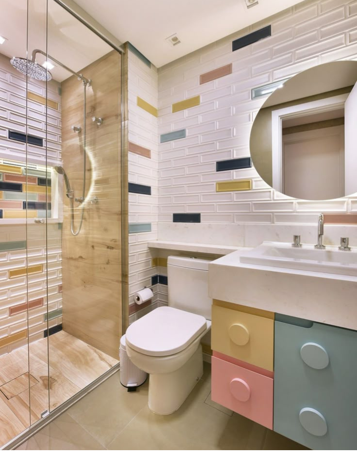 Playful modern bathroom featuring a pastel color-blocked vanity with large round handles, white subway tiles with scattered colored accents, a round backlit mirror, and a glass shower partition with wood-look tile.