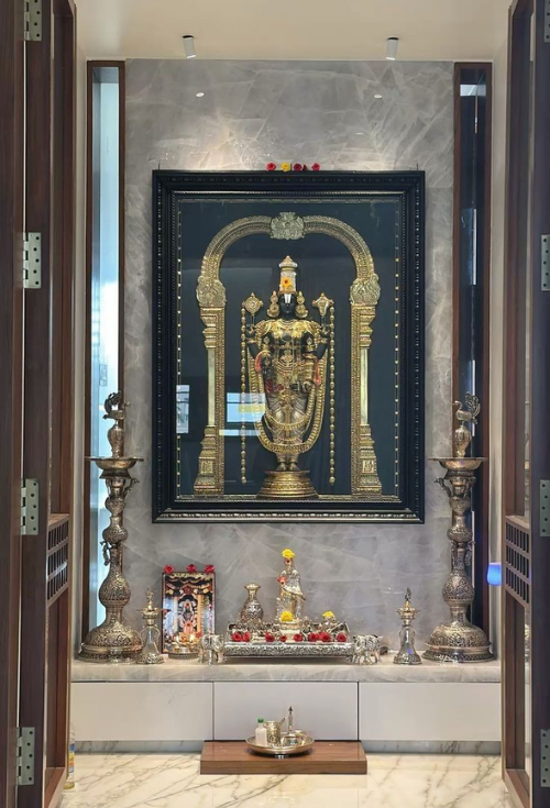 Dedicated Pooja room interior featuring a large framed golden Lord Venkateswara idol against a grey onyx marble backdrop, tall traditional silver lamps, a white marble counter with seamless drawers, and folding wooden doors.