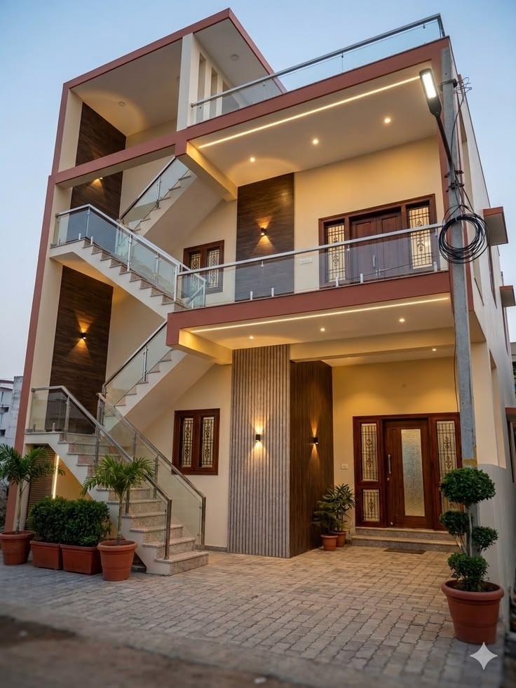 Exterior view of a modern multi-story independent house in Hyderabad featuring glass staircase railings, warm facade lighting, and custom wooden doors.