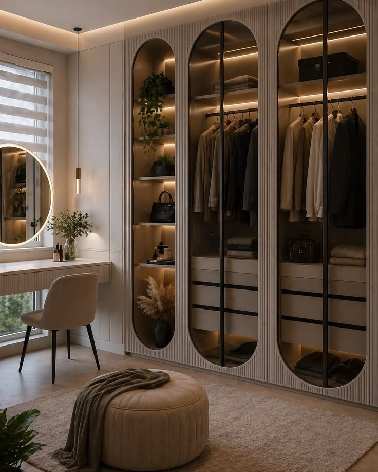 Luxury walk-in wardrobe featuring fluted wall panels, custom capsule-shaped glass display cabinets with integrated LED lighting, a round central pouf, and a floating vanity desk with a backlit round mirror by the window.