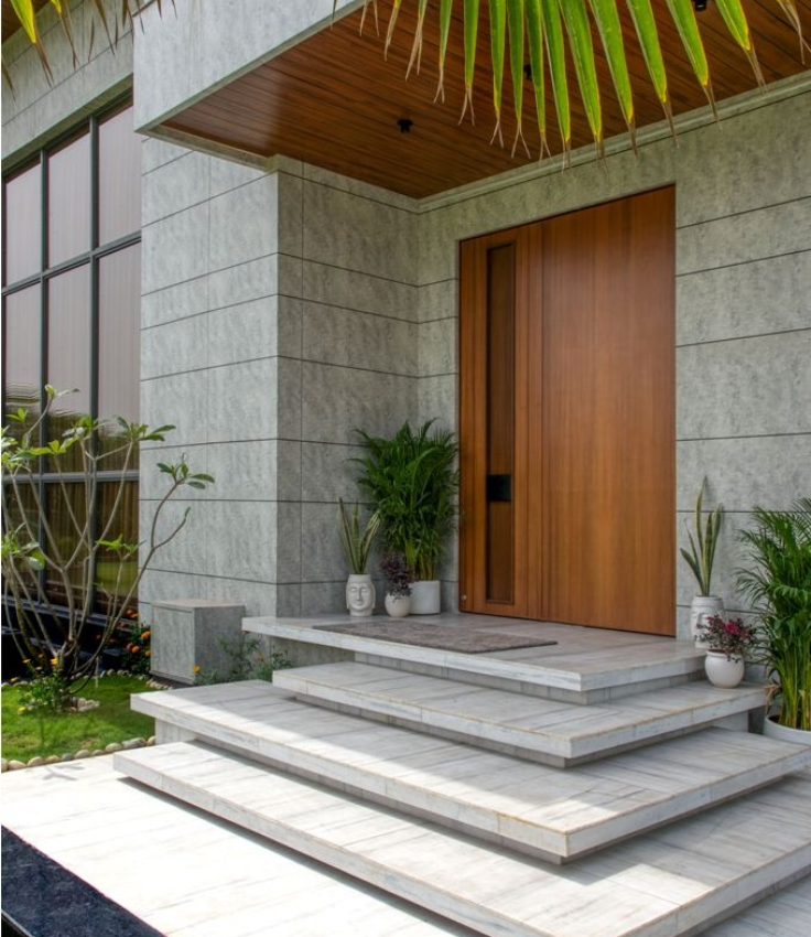 Modern luxury villa main entrance featuring grand floating stone steps, an oversized wooden pivot front door with a vertical glass sidelight, textured grey tile cladding, and a slatted wood porch ceiling.