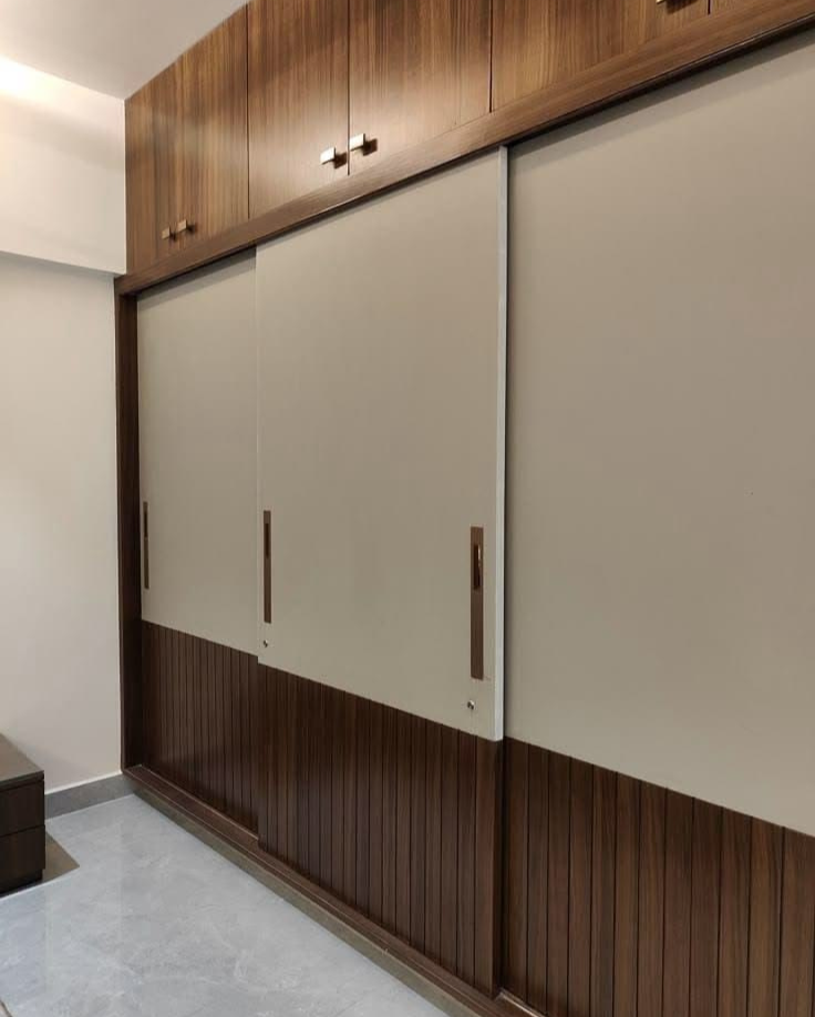 Modern floor-to-ceiling sliding wardrobe featuring a dual-tone finish with matte beige upper panels, fluted wood-grain lower panels, sleek vertical handles, and matching wood-finish top loft cabinets.